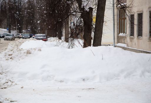 A small snowdrift against the background of a city street with trees. On the road lies white snow in high heaps. Urban winter landscape. Cloudy winter day, soft light.