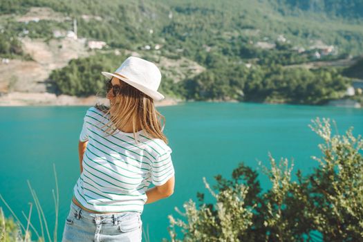 Smiling woman in hat and sunglasses with wild hair standing near mountains lake on background. Positive young woman traveling on blue lake outdoors travel adventure vacation.