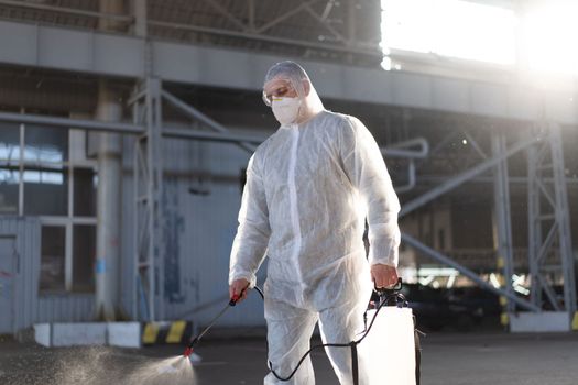 Man dressed white protective overalls spraying surface antibacterial sanitizer sprayer during quarantine Caucasian person protective suit disinfects parking during covid pandemic backlight