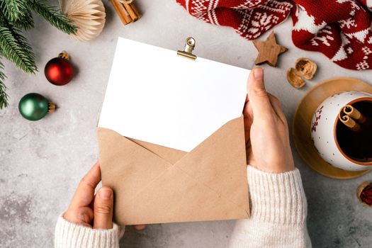 Woman hands in sweater holding blank paper and envelope . Flat lay of gray background with cup of coffee and Christmas decoration. Top view mock up and copy space for text.