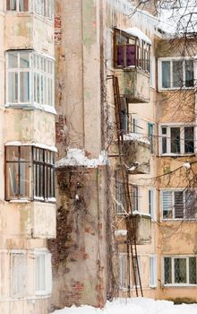 An old four-story house with glazed balconies, a stone chimney on the wall. Plastic windows and an iron ladder on the wall of a shabby house. Cloudy winter day, soft light.