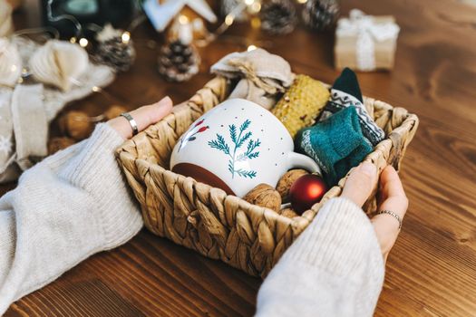 Woman s hands wrapping Christmas eco gift wicker basket, close up. Unprepared presents on wooden table with natural decor elements and items Christmas packing Concept.