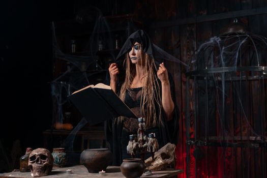 Witch dressed black hood with dreadlocks standing dark dungeon room use magic book for conjuring magic spell. Female necromancer occultist medium shoot