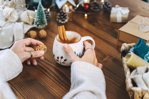 Christmas mug of coffee with cinnamon in woman hands. Wrapping Christmas eco gift box, close up. Preparing presents on wooden table with natural decor elements and items Christmas packing Concept.