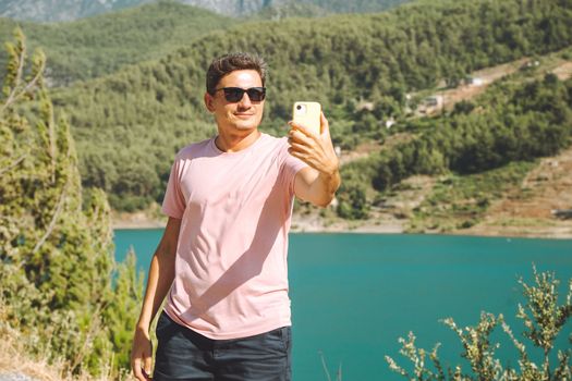 Man taking selfie on mobile phone of mountains lake background. Traveler male having video chat on cellphone on the blue lake outdoors travel adventure vacation.