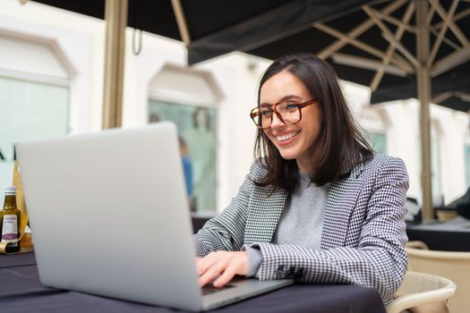 Modern freelancer business woman working remotely at outdoor summer city cafe taking notes use laptop. Caucasian female business person in eyeglasses use laptop sitting street restaurant side view