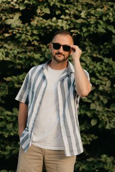 A smiling man is posing in the park in the evening. A happy guy is touching his sunglasses with green leaves in the background.