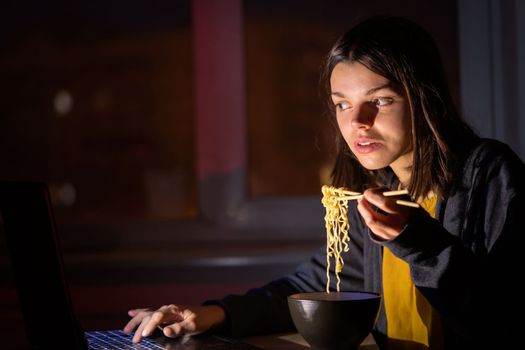 Young girl works at night at home with laptop and eats noodles, asian food with chopsticks. A woman watches a show, a movie and has dinner with ordered food from a cafe.