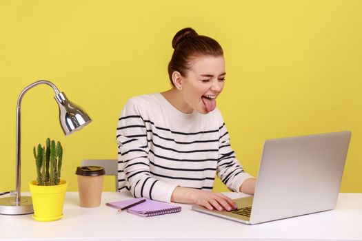 Playful funny woman office worker in striped shirt showing tongue out, expressing positive childish emotions, chatting via web camera on laptop. Indoor studio studio shot isolated on yellow background