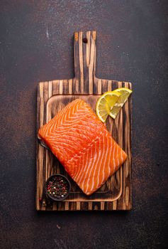 Fresh raw salmon fillet on wooden kitchen cutting board with seasonings and lemon top view on dark brown rustic background