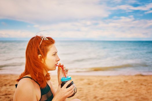 Red-haired girl in a swimsuit is sitting on the beach with a cocktail. Calm woman on vacation. Holidays at the sea. Vintage sunglasses on the head of a having a rest lady.