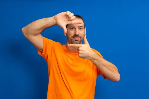 Handsome bearded happy male blinks his eyes and looks through a frame formed by his hands. An attractive man makes a frame with his fingers, isolated on a blue background with copy space for text.