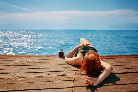 Female in a swimsuit is lying on a wooden pier with a bottle of lemonade and sunbathing. Red-haired woman is enjoying a vacation at the sea. Summer trip. Copy space for travel agency advertising.