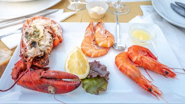 White plate with cooked shrimps, lobster and prawns with herb, sauces and lemon slice.