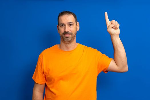 Insightful smart proactive caucasian man 40s in orange t-shirt holding index finger up with great new idea isolated on blue background studio portrait. People lifestyle concept.