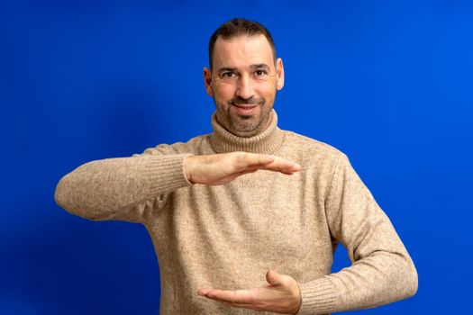 Happy hispanic man wearing a brown turtleneck showing something between his hands, product marketing advertisement concept with copy space over blue background