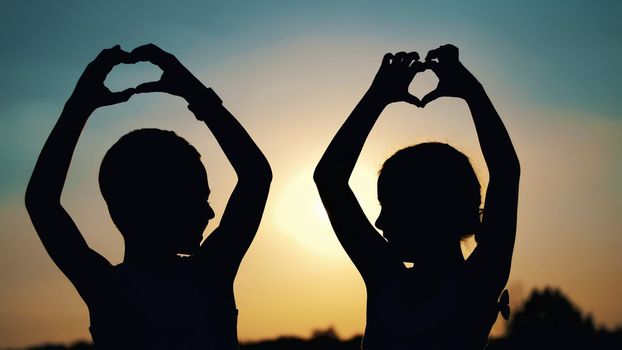 Silhouettes, figures of children, boy and girl show the hearts with the fingers, hands, against the background of the sun, at sunset in summer. Happy family. Slow motion. High quality photo