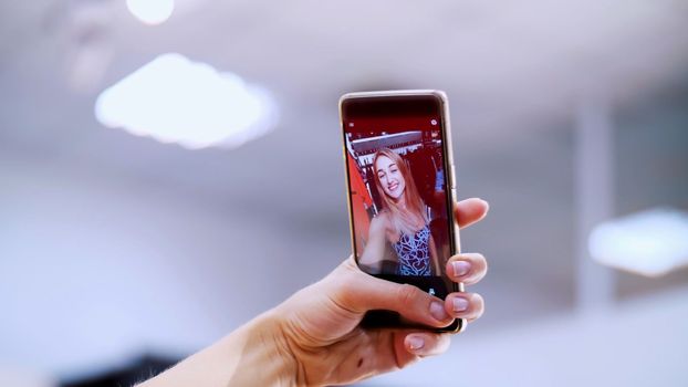 Sexy girl, a beautiful blonde woman makes selfie in a new outfit, smiling, in a store, a boutique of clothes. In the frame is a female hand with a smartphone. slow motion. High quality photo