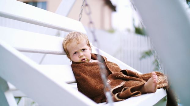 a little one-year-old girl, wrapped in a towel, with a dirty face, lonely sitting on a swing in the garden, in the summer. She has a sad look. She wants to sleep. High quality photo