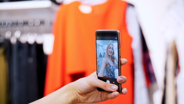 Sexy girl, a beautiful blonde woman makes selfie in a new outfit, smiling, in a store, a boutique of clothes. In the frame is a female hand with a smartphone. slow motion. High quality photo