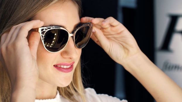 Sexy girl, tall, beautiful blond woman trying on sunglasses in a stylish store, boutique and admiring herself in front of a mirror, smiling. slow motion. close - up. High quality photo