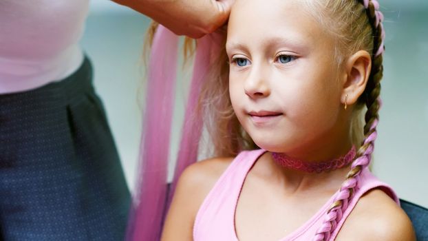 Beautiful blond girl, of seven years old, braided two pigtails, do a hairstyle with pink locks of hair in a beauty salon, a hairdresser's salon, in front of a large mirror. a little princess. High quality photo