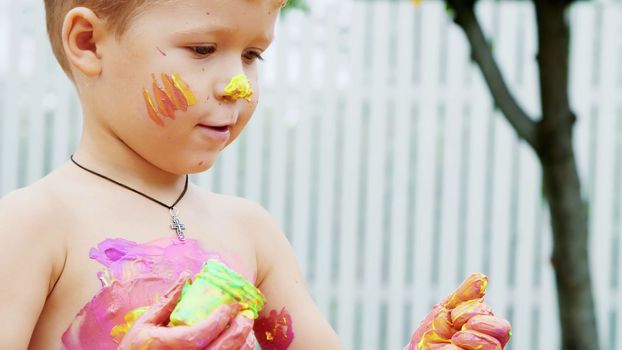 a little child, a four-year-old boy playing, painting with finger paints, decorating himself, in the garden, sitting on a blanket, on grass, lawn, in the summer. he's having fun. High quality photo