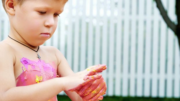 a little child, a four-year-old boy playing, painting with finger paints, decorating himself, in the garden, sitting on a blanket, on grass, lawn, in the summer. he's having fun. High quality photo