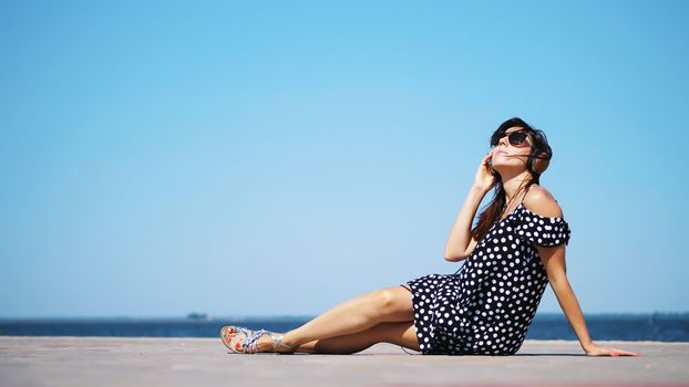 beautiful brunette girl in sun glasses and sundress, with big headphones, listening to music from smartphone, on beach, on hot summer day, near sea. High quality photo