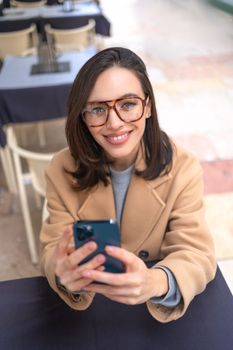 Woman using smartphone sitting street cafe. portrait modern caucasian woman in glasses holding mobile phone in hand looking at camera. Vertical photo. Positive smiling female person with smartphone