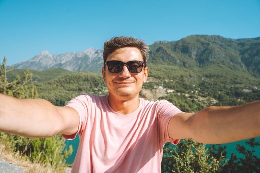 Man taking selfie photo with wide angle front camera of mountains lake background. Traveler male having fun on the blue lake outdoors travel adventure vacation.