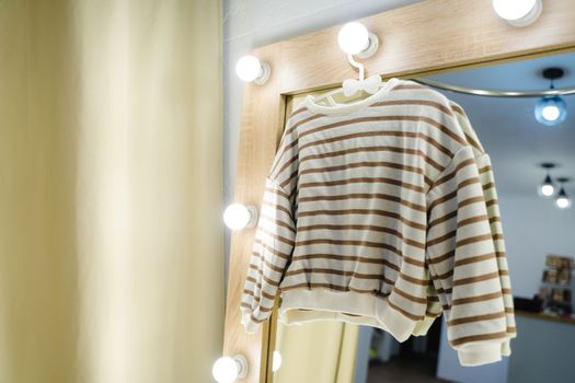 Jumper hangs on a hanger on a mirror with light bulbs. Fitting room with a large beautiful mirror with light bulbs. Children's sweater with stripes. Beige sweater with brown stripes.
