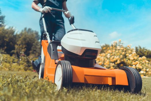 A young man in a straw hat is mowing a lawn with a lawn mower in his beautiful green floral summer garden. A professional gardener with a lawnmower cares for the grass in the backyard.