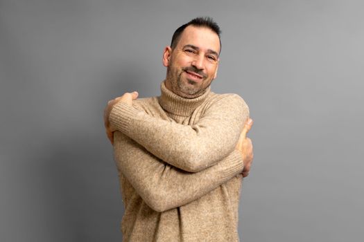 Portrait of egotistical bearded dark-haired man in turtleneck sweater with closed eyes, hugging himself and smiling from pleasure and pride, isolated over gray background