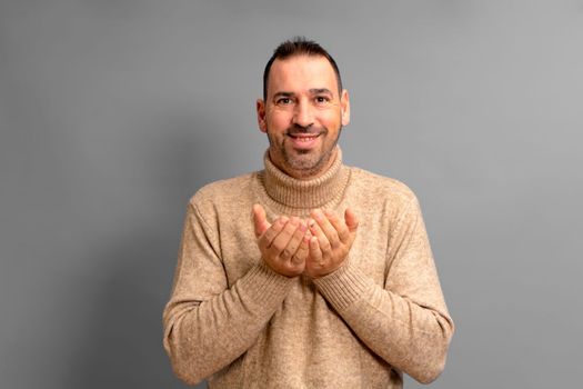 Bearded latin man dressed in a turtleneck sweater offering something imaginary in his hands close to the chest isolated on a gray studio background, advertising concept, copy space
