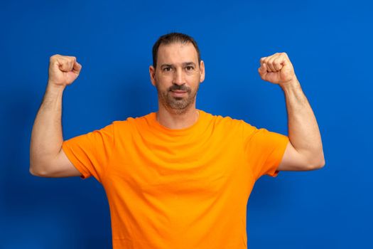 Hispanic male happy unshaven strong sporty fitness student 40s man in orange t-shirt showing biceps muscles on hand isolated on blue color background studio portrait