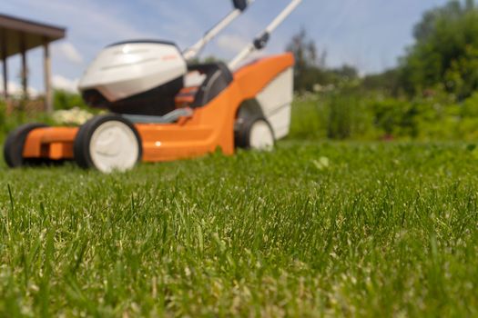 A lawn mower is outside at the beautiful green floral backyard lawn. A lawnmower is cutting a lawn on a summer sunny day.