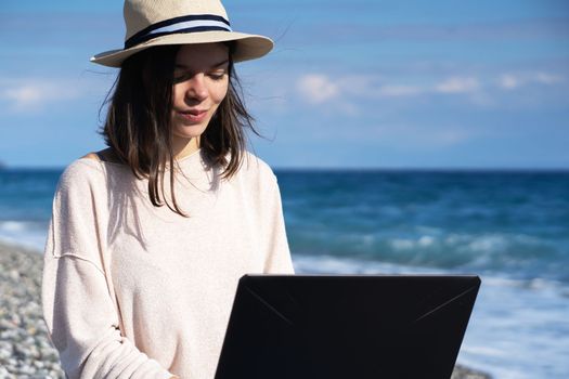 A young smiley girl in a hat and casual sweater sits on the beach by the sea with a laptop on a sunny day, works, studies, buys tickets during trip, woman rests on vacation and types on the keyboard.