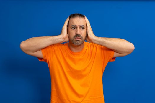 Stunned latin man wearing an orange t-shirt with his head in his hands isolated on a blue studio background