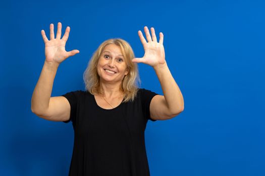 Pretty caucasian blonde woman in her 40s dressed in a black t-shirt raising her palms up while smiling at the camera, isolated on blue studio background