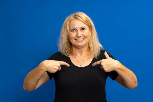 Portrait of a charming happy woman in a black t-shirt smiling and pointing at herself, proud of her own success, bragging about her achievements. Indoor studio shot isolated on blue background.