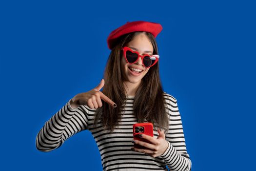 Smiling teenager girl pointing to smartphone, she advise mobile app, mobile operator. Young woman on blue background. . High quality photo