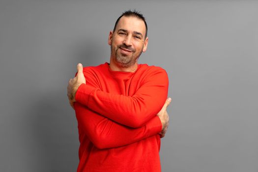 I love myself. Portrait of an egotistical bearded dark-haired man in a red pullover with his eyes closed, hugging himself and smiling with pleasure and pride. Indoor, isolated on gray background