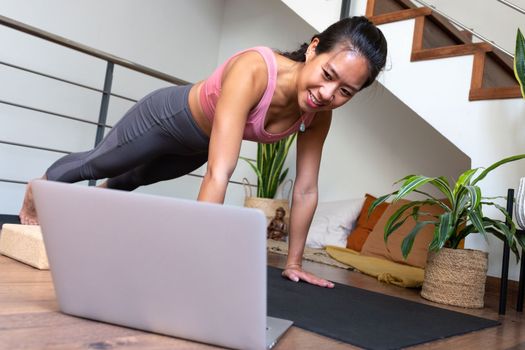 Chinese young woman doing core strengthening exercises at home. Asian female doing plank pose using online video training with laptop. Active and healthy lifestyle concept.