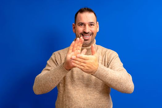 Bearded hispanic man wearing brown turtleneck sweater over isolated blue studio background clapping hands happy and cheerful, smiling proudly with hands together