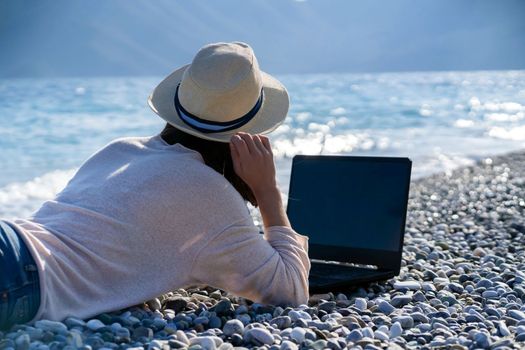 A young girl in a light hat and casual sweater lies on the beach by the sea with a laptop on a sunny day, works, studies, buys tickets during trip, a woman rests on vacation and types on the keyboard.