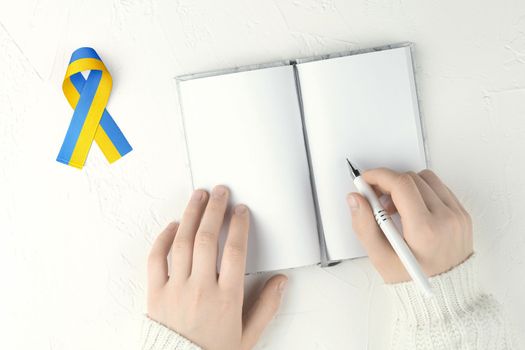 female hands with pen an open blank notepad and yellow blue ribbon on white table. concept needs help and support, truth will win