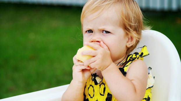 summer, in the garden. portrait, one year old pretty blond girl eating an apple with apetite. on the table. High quality photo