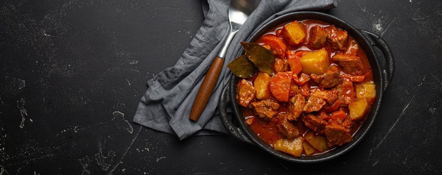 Beef meat stew with potatoes, carrot and delicious gravy in black casserole pot with bay leaves with spoon on black dark rustic concrete background from above, space for text.