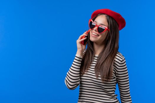 Stylish french woman talking by phone, nice joke, smiling. Young lady on blue background. Having smartphone call. . High quality photo